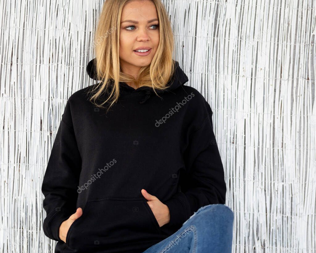 Sweatshirt hoodie mockup. A nice smiling blonde poses in a black sweatshirt against a white wall. She is sitting facing the camera.