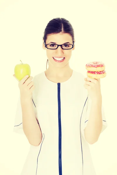 Young female doctor, nurse holding an apple and doughtnuts. — Stock Photo, Image