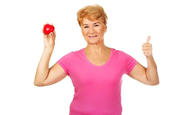 Viejo sonriente mujer sosteniendo rojo juguete corazón con pulgar hacia arriba — Foto de Stock