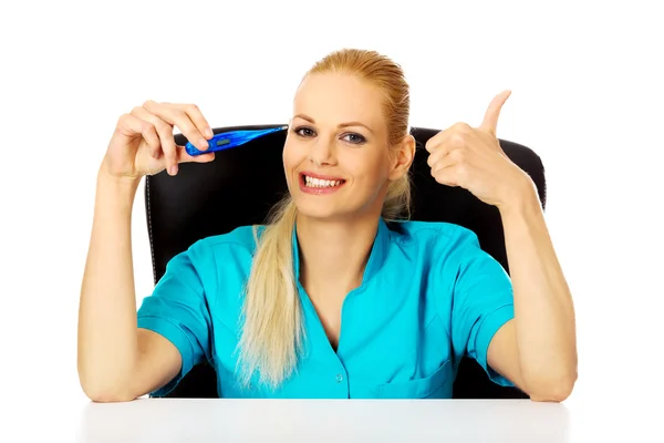 Médico o enfermera sonriente sentada detrás del escritorio sosteniendo el termómetro y mostrando el pulgar hacia arriba —  Fotos de Stock