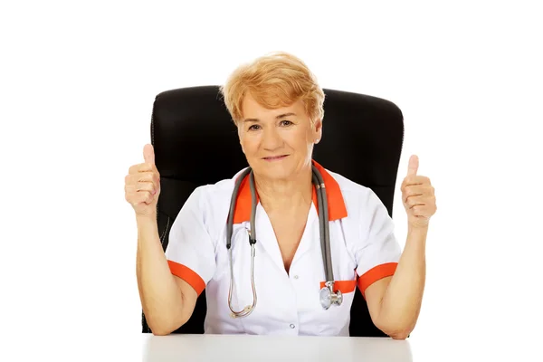 Улыбается пожилая женщина врач или медсестра сидит за столом и показывает большие пальцы вверх — стоковое фото