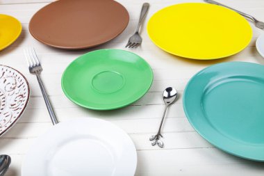 Ahşap zemin üzerinde çok renkli tabaklar, kaşıklar ve çatallar boş, üst görünüm.