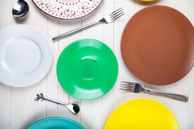 Ahşap zemin üzerinde çok renkli tabaklar, kaşıklar ve çatallar boş, üst görünüm.