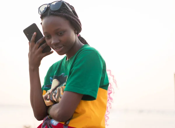 Young beautiful black girl using smarphone on sea beach at sunset Лицензионные Стоковые Фото