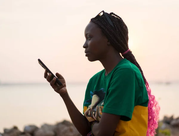 Молода красива чорна дівчина використовує смартфон на морському пляжі на заході сонця Стокове Фото