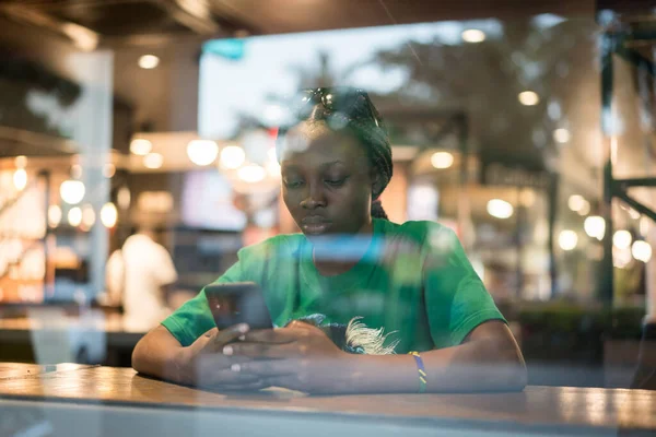 Young authentic black woman sitting with phone in city coffee shop at night