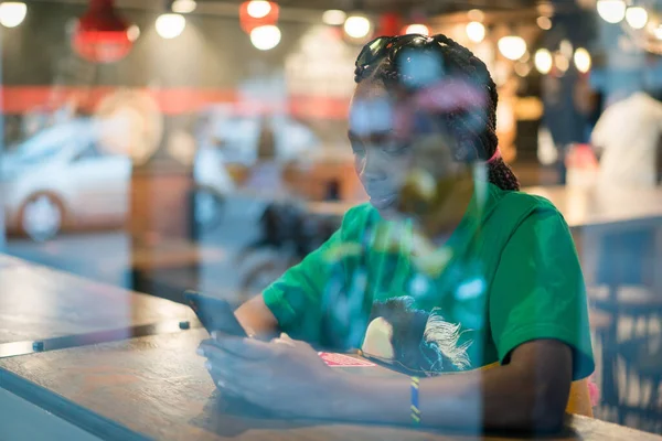 Young authentic black woman sitting with phone in city coffee shop at night