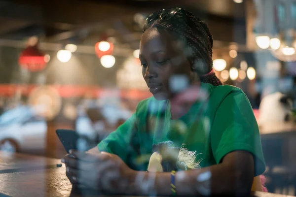 Junge authentische schwarze Frau sitzt nachts mit Telefon im städtischen Café Stockbild