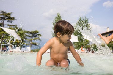 Küçük çocuk yüzme havuzunda su sıçratıyor, eğlenceli aktiviteler yapıyor.