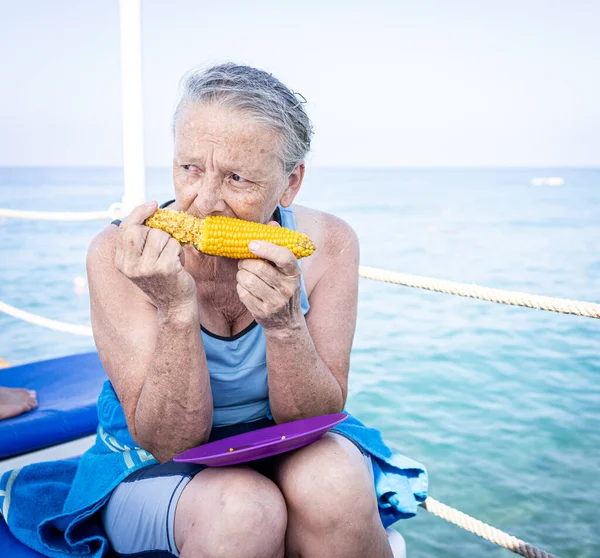 Ανώτερη γυναίκα κάθεται στην προβλήτα παραλία και τρώει καλαμπόκι — Φωτογραφία Αρχείου