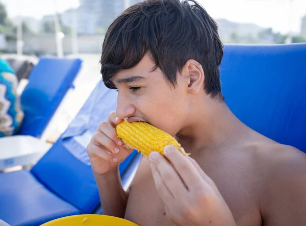 Jongen zittend op strand pier en het eten van maïs — Stockfoto