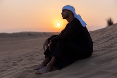 Arap adam altın saat boyunca çölde kumların üzerinde oturuyor, geleneksel Arap kıyafetleri giyiyor. Yüksek kalite fotoğraf
