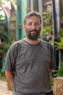 Sakallı ve gri gömlekli bir adam bir binanın önünde duruyor. Ciddi görünüyor ve bir şeye bakıyor.