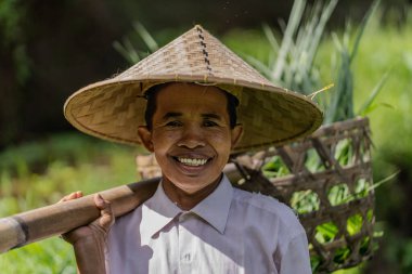 Hasır şapka takan bir kadın gülümsüyor ve elinde bir sepet tutuyor. O yemyeşil bir arazide.