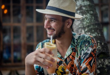 Tropikal tişörtlü bir adam elinde pipetle içki tutarken gülümsüyor. İçki bir limonatadır ve adamın hoşuna gider.