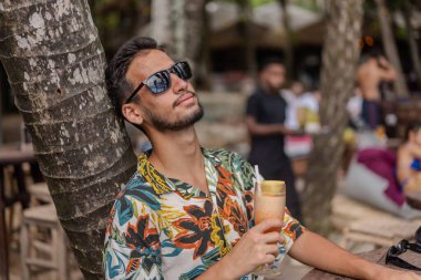 Sakallı ve gri gömlekli bir adam gülümsüyor ve dirseğine yaslanıyor. O bir saat takıyor ve rahat bir ifadesi var.