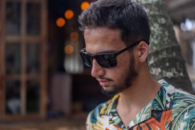 Hawai gömlekli bir adam pipetle içki içiyor. Bir ağacın altında oturuyor.