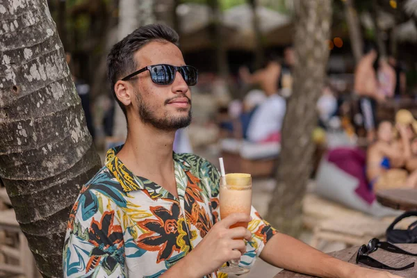 Sakallı ve gri gömlekli bir adam gülümsüyor ve dirseğine yaslanıyor. O bir saat takıyor ve rahat bir ifadesi var.