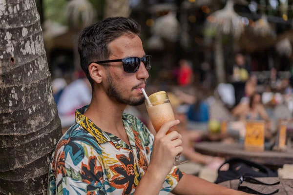 Hawai gömlekli bir adam pipetle içki içiyor. Bir ağacın altında oturuyor.