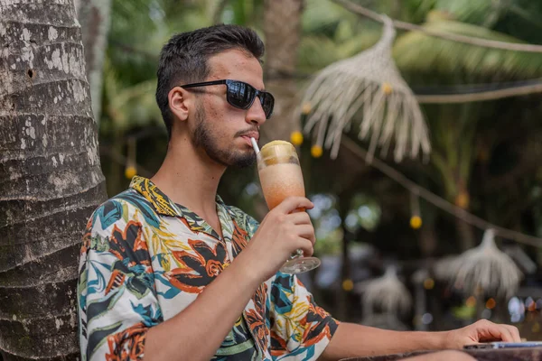 Sakallı ve gri gömlekli bir adam gülümsüyor ve dirseğine yaslanıyor. O bir saat takıyor ve rahat bir ifadesi var.