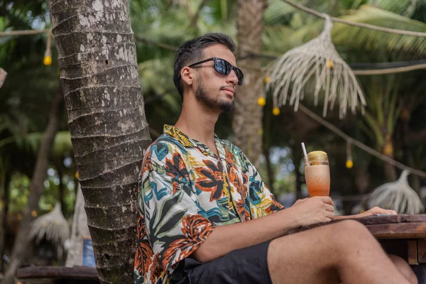 Sakallı ve gri gömlekli bir adam gülümsüyor ve dirseğine yaslanıyor. O bir saat takıyor ve rahat bir ifadesi var.