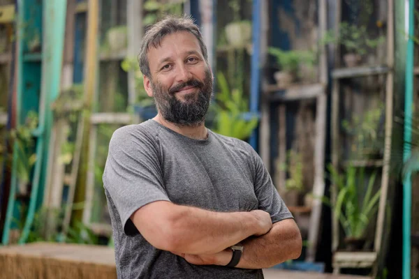 Sakallı ve gri gömlekli bir adam bir binanın önünde duruyor. Ciddi görünüyor ve bir şeye bakıyor.