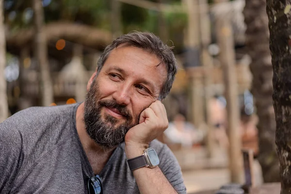 Sakallı ve gri gömlekli bir adam gülümsüyor ve dirseğine yaslanıyor. O bir saat takıyor ve rahat bir ifadesi var.