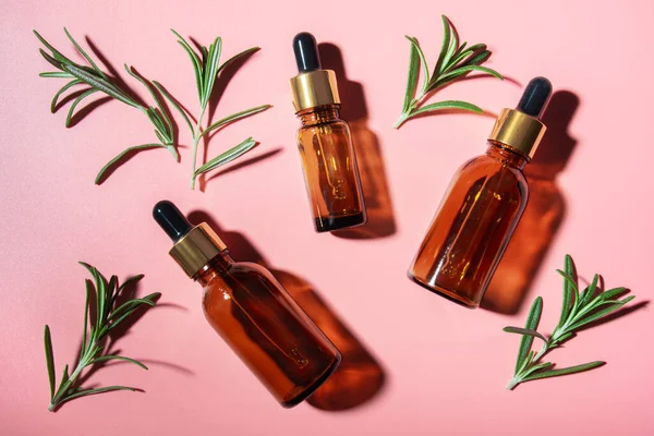 Dropper bottles with oil and rosemary leaves on pink table flat lay view. Herbal cosmetics concept