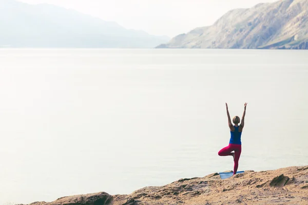 Woman meditating in yoga tree pose at the sea and mountains — Stok fotoğraf