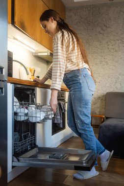 Kadın bulaşık makinesini açıyor. Zaman kazandıran teknoloji. Dahili bulaşık makinesini aç. Entegre aletleri olan beyaz mutfak. Bulaşık makinesinde temiz tabaklar ve bulaşıklar
