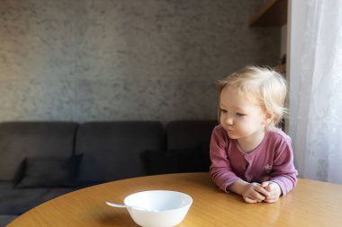 Aç küçük kız sandalyeye oturur ve kaşıkla yulaf lapası yer. Bulanık yemek odası arka planı. Çocuklar için sağlıklı beslenme. Çocuk beslenmesi