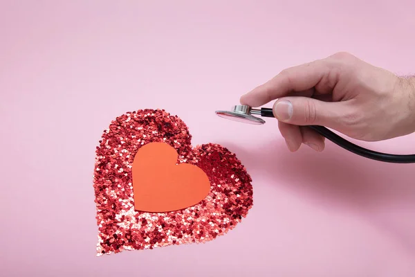 La mano del médico con un statoscopio escucha el corazón hecho de muchas lentejuelas rizadas en forma de corazones —  Fotos de Stock