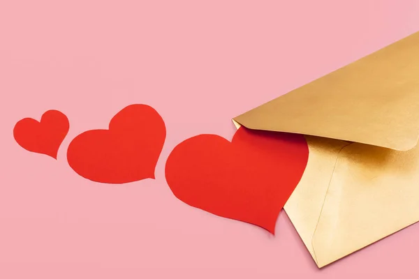 Золотий паперовий конверт з червоними серцями біля нього ізольований на рожевому фоні — стокове фото