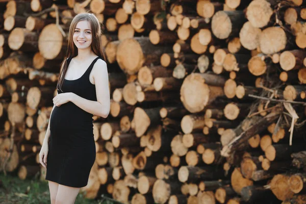 一个穿着黑色衣服的年轻貌美的怀孕少女站在大自然的背景上 背靠折叠的圆木支撑着她的肚子 强调它是圆形的 — 图库照片