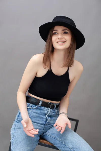 Stüdyo portresi, gri arka planda siyah tişörtlü, mavi kot pantolonlu ve sandalyede oturan esmer bir kızın resmi.