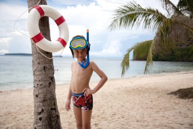 Dalgıç maskeli bir çocuk sahilde palmiye ağacının yanında duruyor.