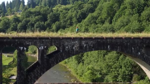 明るい夏の日に古い石造りの橋を渡って山の川を渡るハイカーの女の子 — ストック動画
