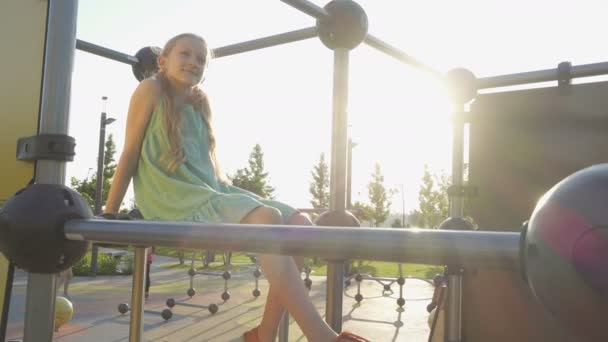 小女孩坐在早上的操场上 在升起的阳光下摆动着双腿 — 图库视频影像