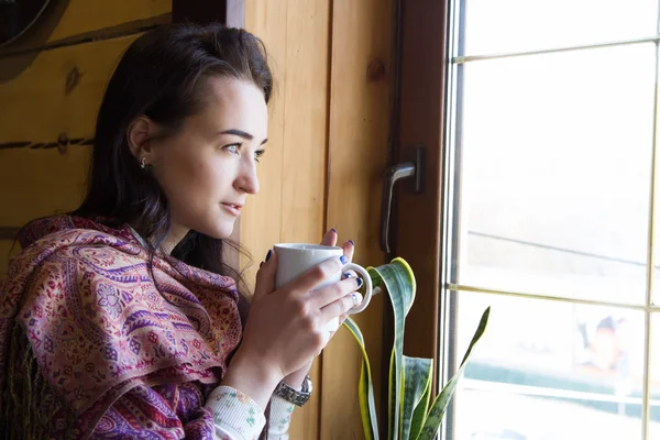 Chica joven con una taza de café de pie en la ventana — Foto de Stock