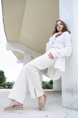 İnce bir genç kız bir binanın sütununa yaslanmış duruyor.