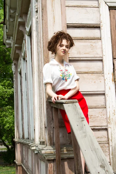 Young girl in a white embroidered shirt standing on the porch of a wooden house — Stock Photo, Image