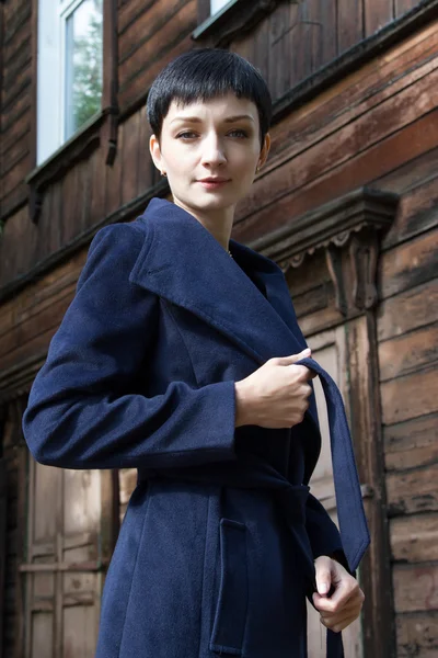 Menina nova em um casaco em um contexto de uma velha casa de madeira — Fotografia de Stock
