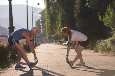 Koşan çift sabah olmadan ısınıyor ve esniyor. Arka planda gündoğumuyla birlikte şehirde antrenman yapıyorlar.