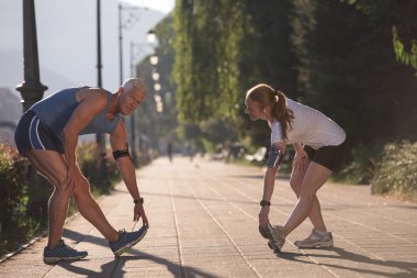 Koşan çift sabah olmadan ısınıyor ve esniyor. Arka planda gündoğumuyla birlikte şehirde antrenman yapıyorlar.
