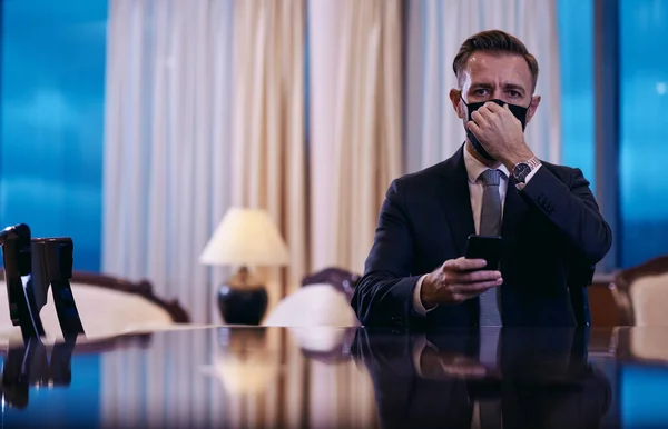 Homem de negócios usando telefone inteligente no escritório de luxo usando máscara facial — Fotografia de Stock