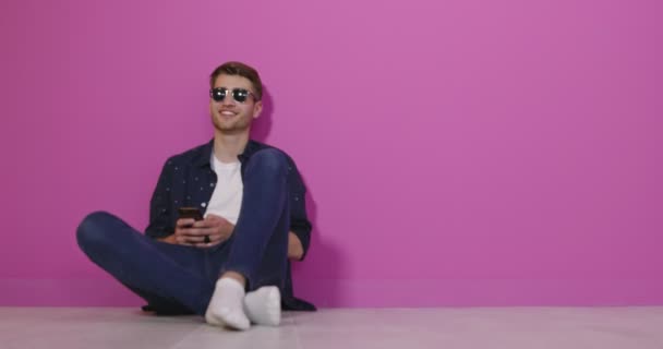 Joven sonriente sosteniendo el teléfono inteligente y mirándolo. Retrato de un hombre feliz usando teléfono móvil aislado sobre fondo rosa. — Vídeos de Stock