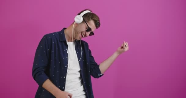 Junger gutaussehender Mann mit Kopfhörern, der Musik über isoliertem Farbhintergrund hört, tanzt und mit erhobenen Armen lächelt — Stockvideo