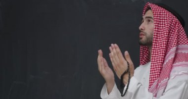  Siyah karatahta önünde dua eden geleneksel kıyafetli genç bir Arap. 
