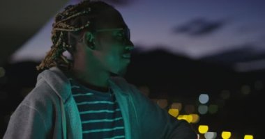 Afro-Amerikalı adam parti veriyor, içki içiyor ve sigara içiyor. Arka planda şehir ışıkları ve neon ışıkları var. 