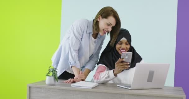 不同种族的妇女在办公室相遇。在Hijab的微笑女孩在笔记本电脑上工作，金发女孩看着屏幕. — 图库视频影像